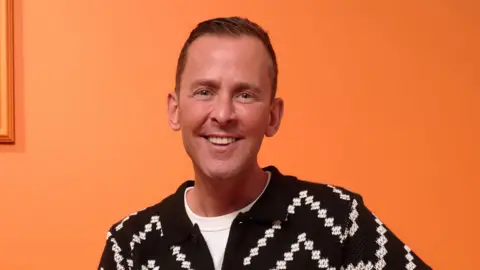 Scott Mills in front of an orange wall. He is sitting down smiling in a black and white shirt.